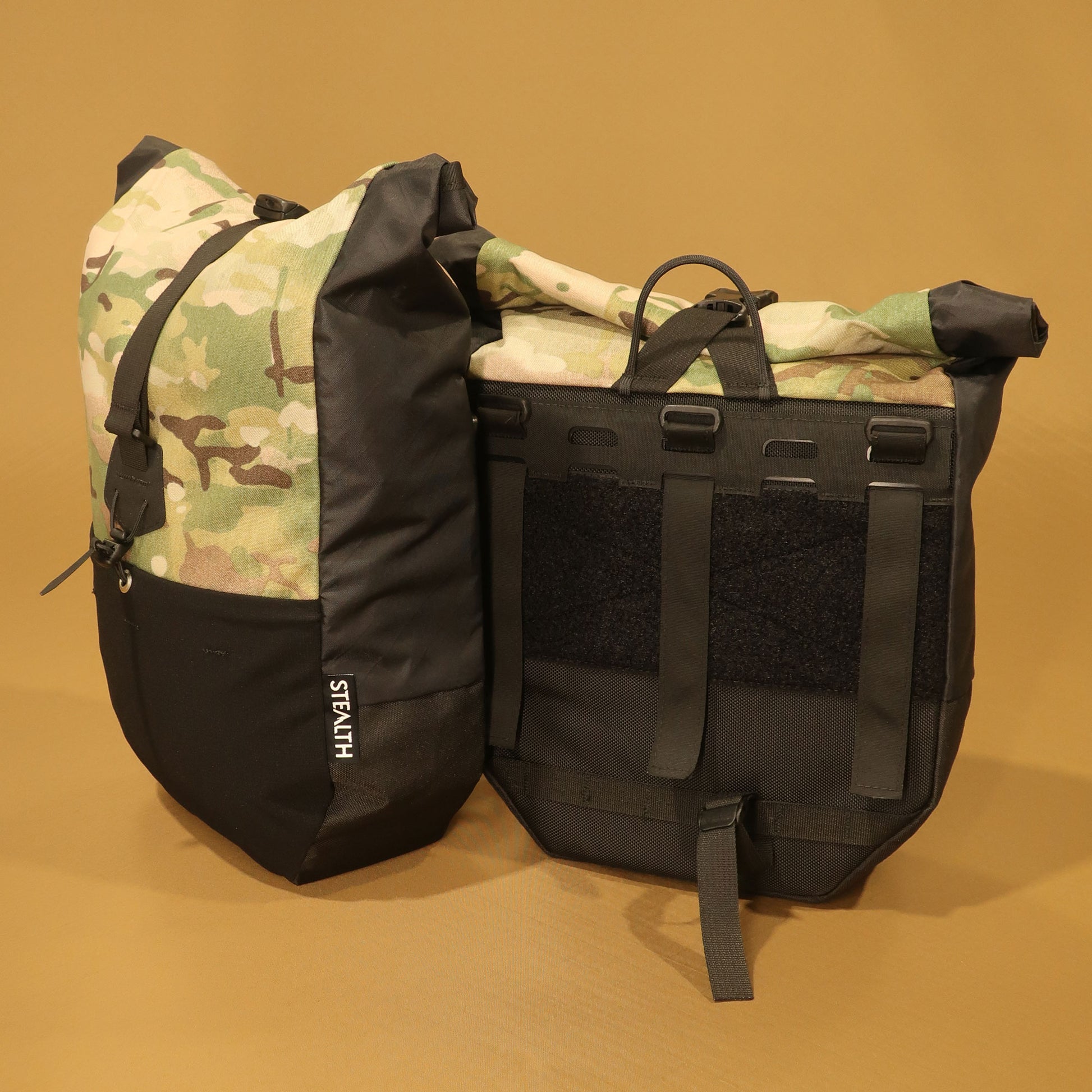 Black and Multicam mountain pannier with a roll top closure and laser cut attachment panel, designed for mountain biking and bikepacking.