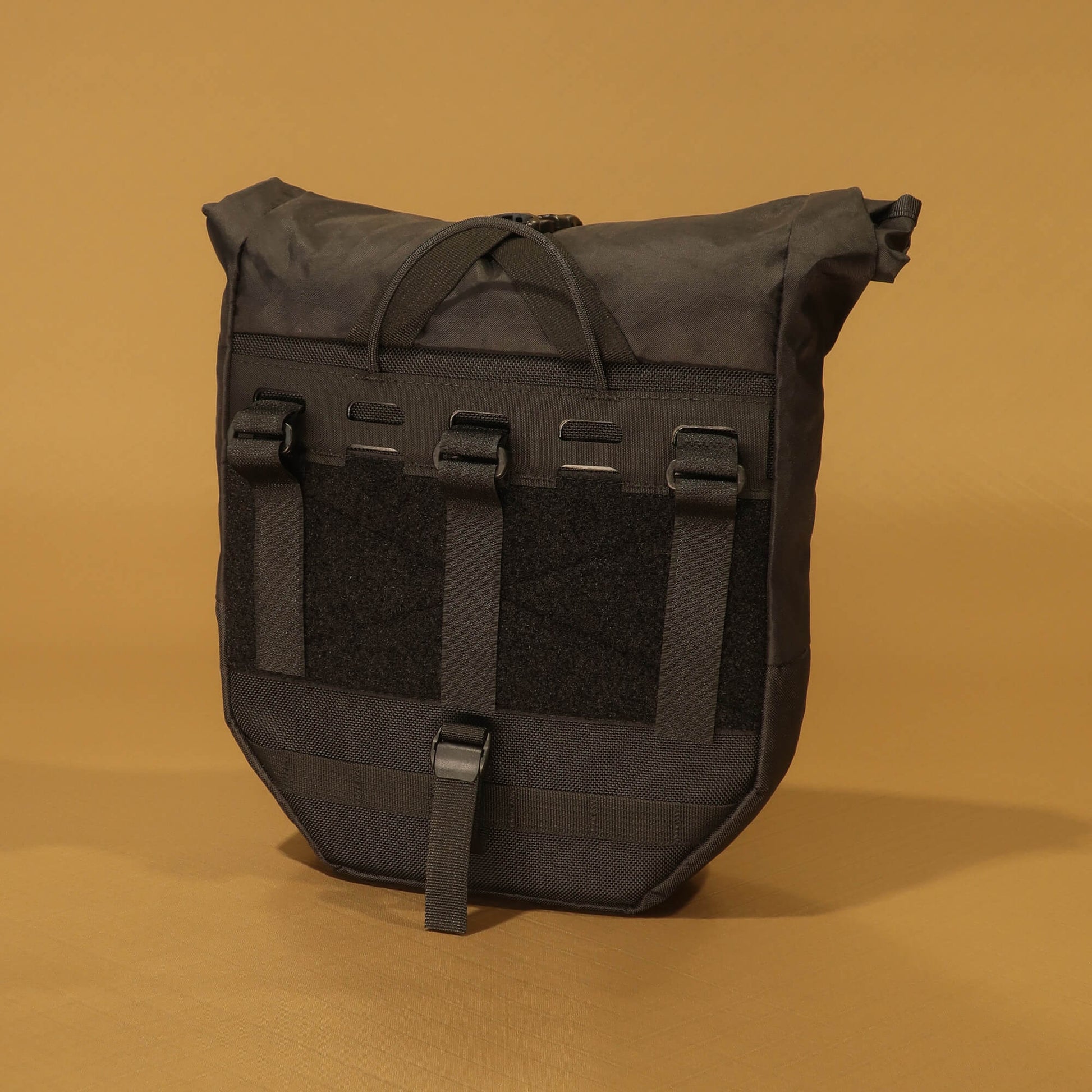 Black roll-top bicycle pannier showing rear rack attachment panel with three hook-and-loop straps, velcro patch, and bottom buckle clip