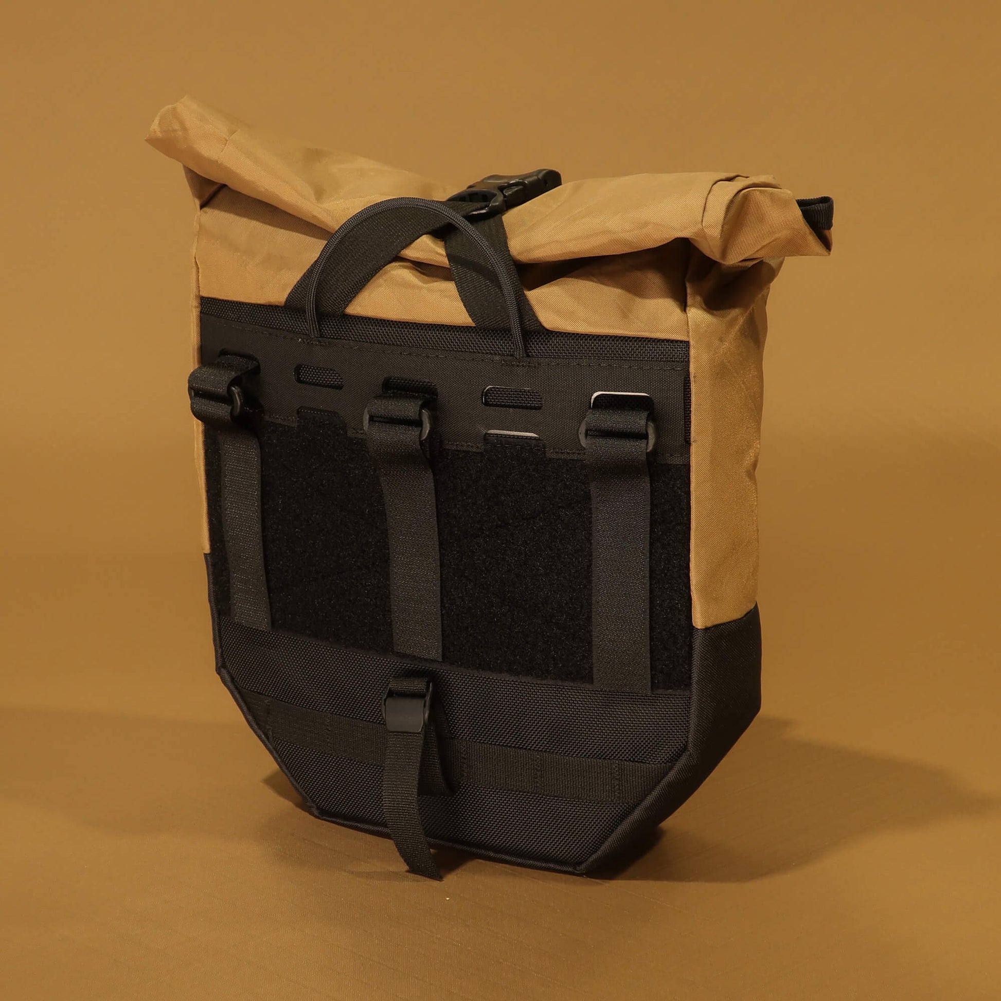 Rear view of 8L roll-top bicycle pannier in coyote tan and black, showing rack attachment panel with hook-and-loop straps, velcro patch, carry handle, and bottom buckle clip