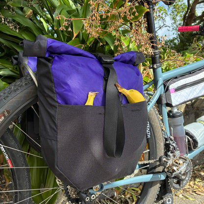 Purple and black roll-top bicycle pannier mounted on a teal cargo bike's rear rack, with two bananas stored in the front pocket, photographed outdoors against lush green foliage